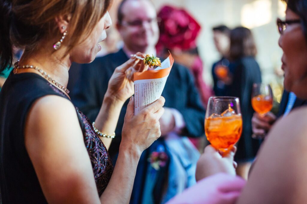 Guests at an event enjoying appetizers and orange cocktails, with a woman holding a paper cone of food in the foreground.