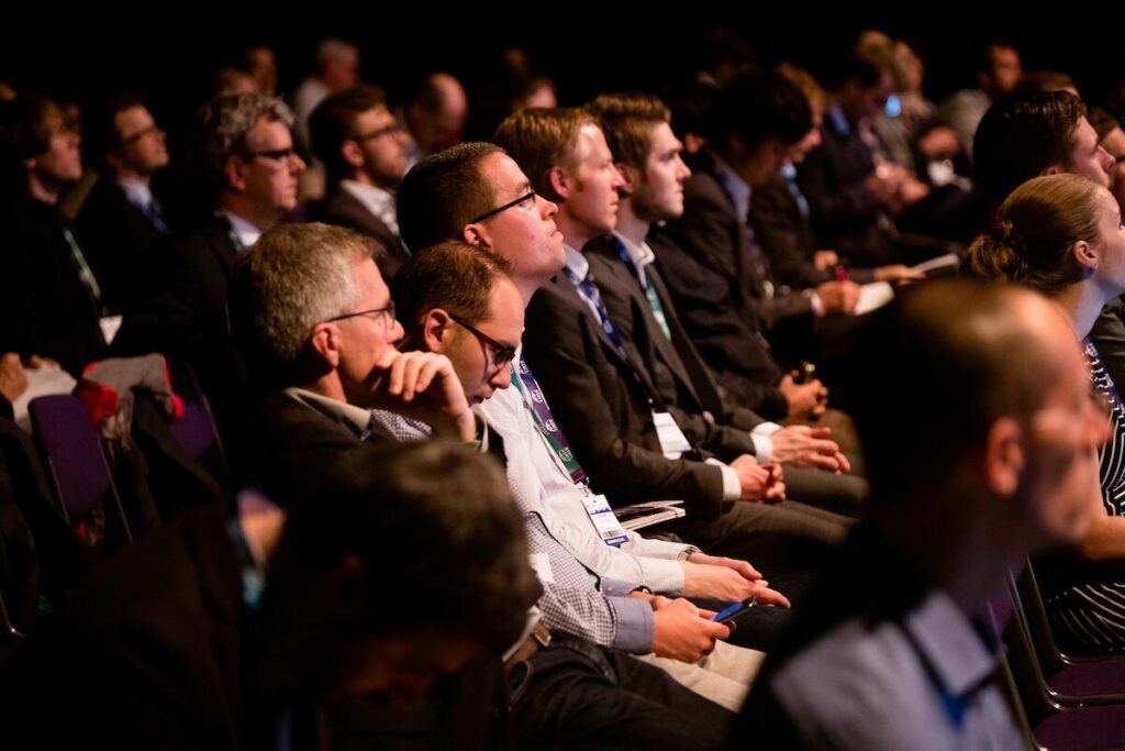 Audience attentively listening to a presentation at a conference, with rows of seated individuals in business attire.