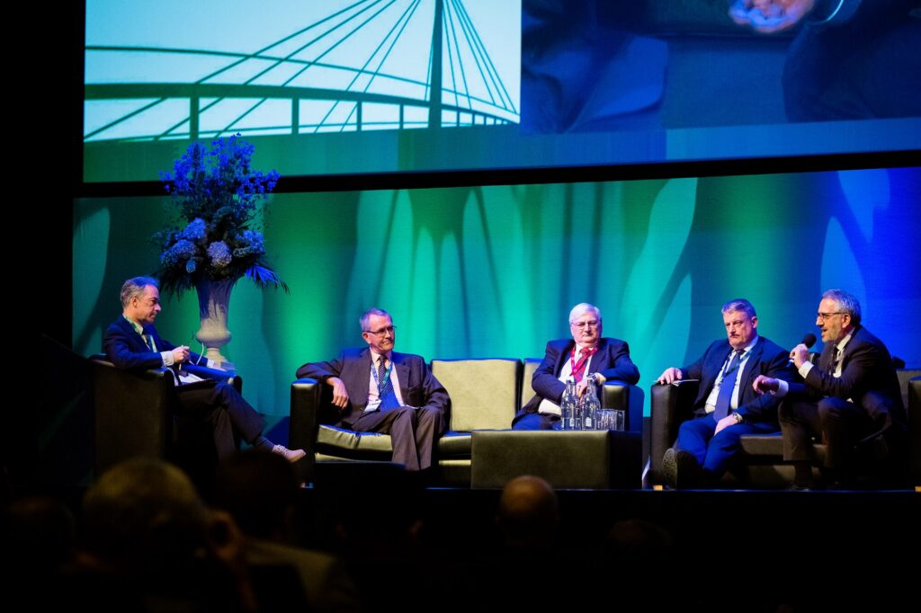 Five men in suits seated on a stage during a panel discussion, with a green and blue backdrop and a large screen above them.