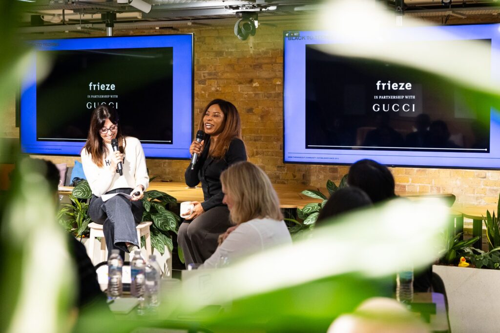 A panel discussion with two speakers, one holding a microphone, seated in front of two screens displaying the logos of Frieze and Gucci, observed by an audience in a room with plants.