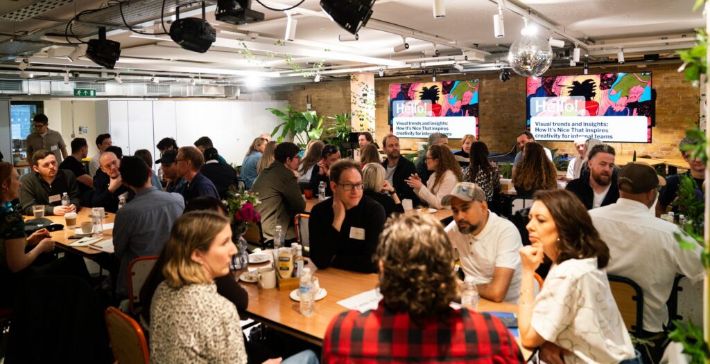 A lively group of people gathered around tables in a modern, well-lit room, engaging in discussions. Two colorful presentation screens are visible in the background.