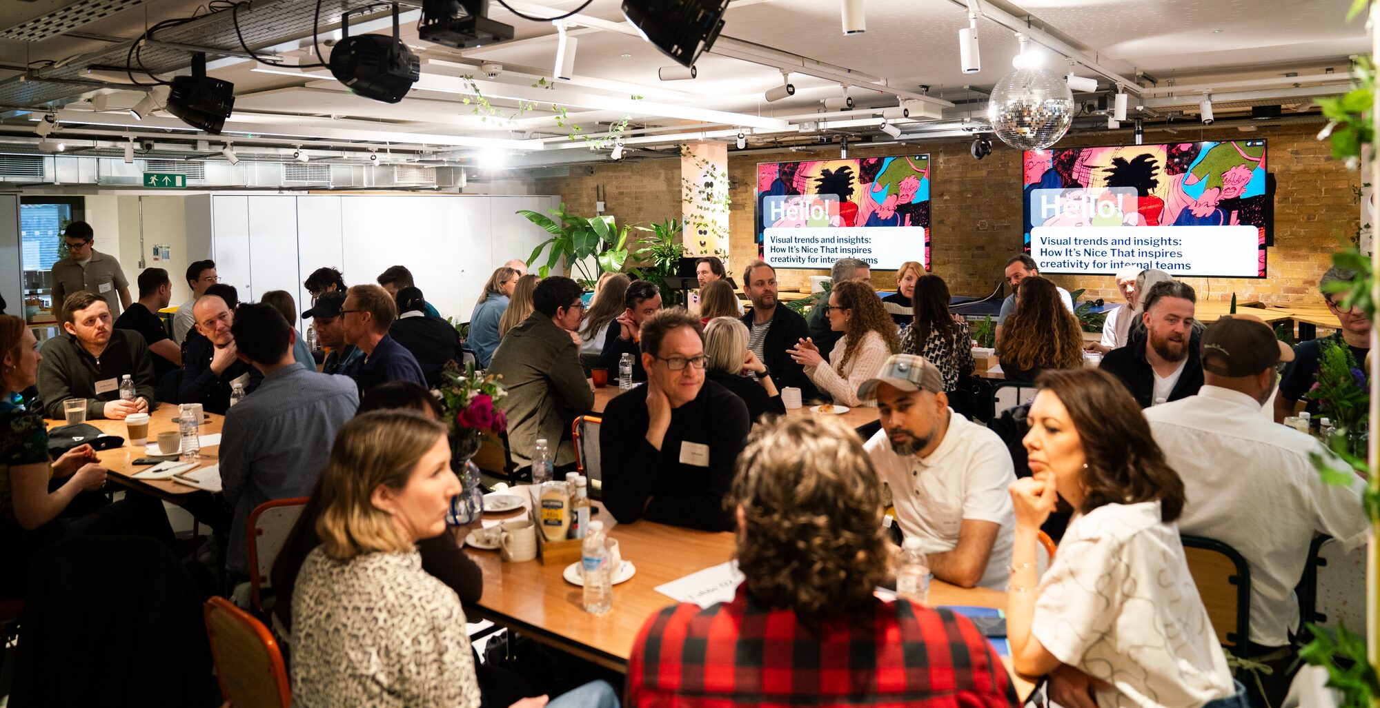 A lively group of people gathered around tables in a modern, well-lit room, engaging in discussions. Two colorful presentation screens are visible in the background.